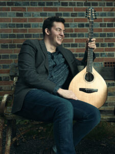 London Irish guitarist and trad musician Tad Sargent smiling with a bouzouki in front of a brick wall