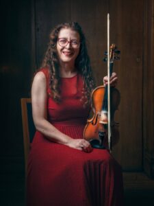 London Irish fiddle player musician Karen Ryan with long curly hair in a red dress smiling holding a fiddle