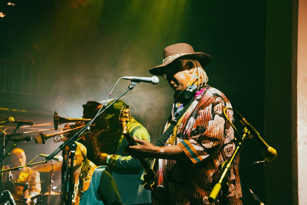 Greg Kofi Brown on stage paying guitar with a brass band in the background